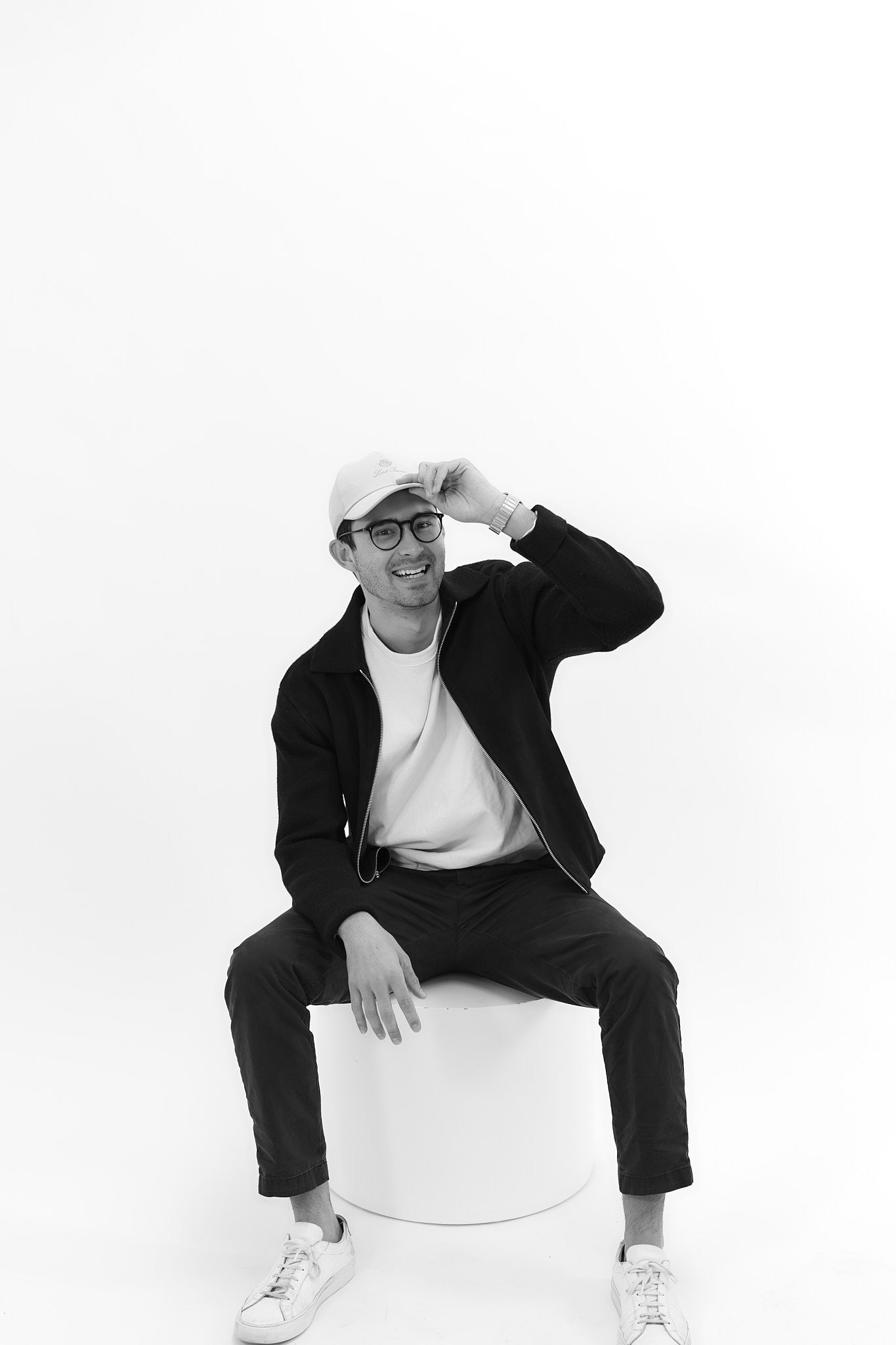 Black and white photo of male in white hat, black glasses, white shirt, black jacket, black pants, white shoes. His left hand is positioned on top of his hat