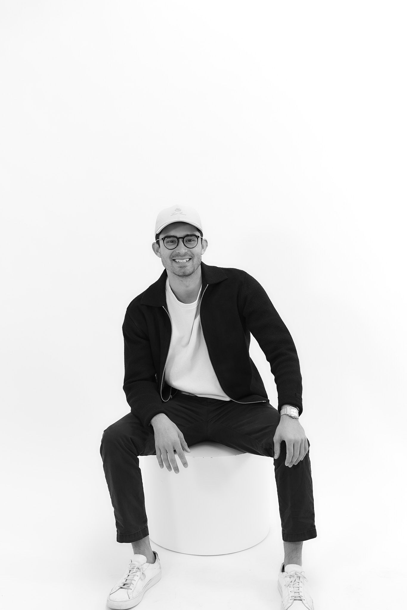 Black and white photo of male in white hat, black glasses, black jacket, black pants, white shirt, white shoes. He is smiling in a seated pose with one arm resting on leg and other arm resting on knee