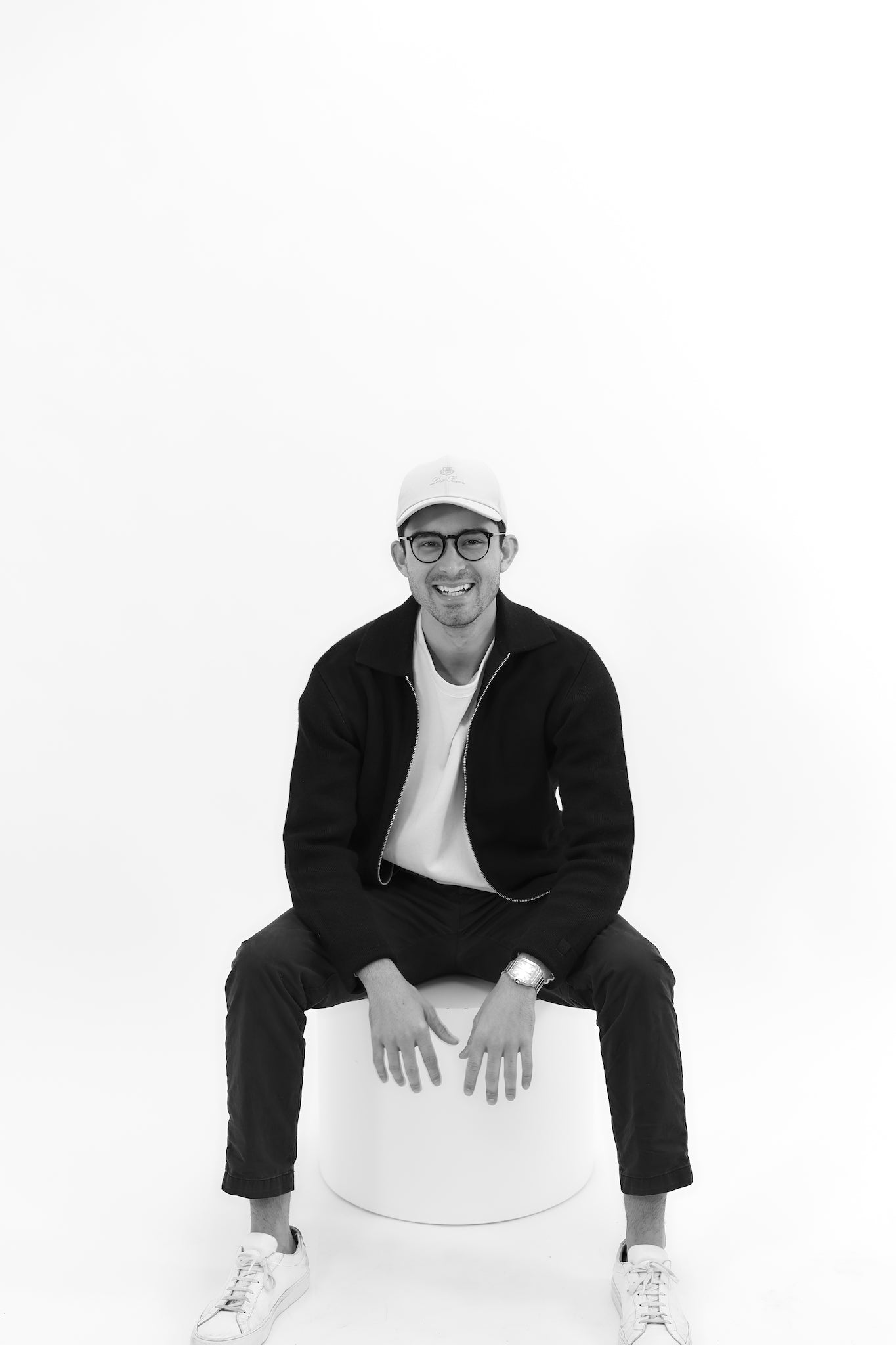 Black and white photo of male in white hat, black glasses, black jacket, black pants, white shirt, white shoes. He is smiling in a seated pose with both arms resting on legs